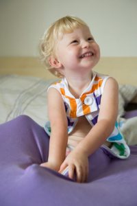 Blond haired toddler sat up in bed in cosi bed sheet smiling