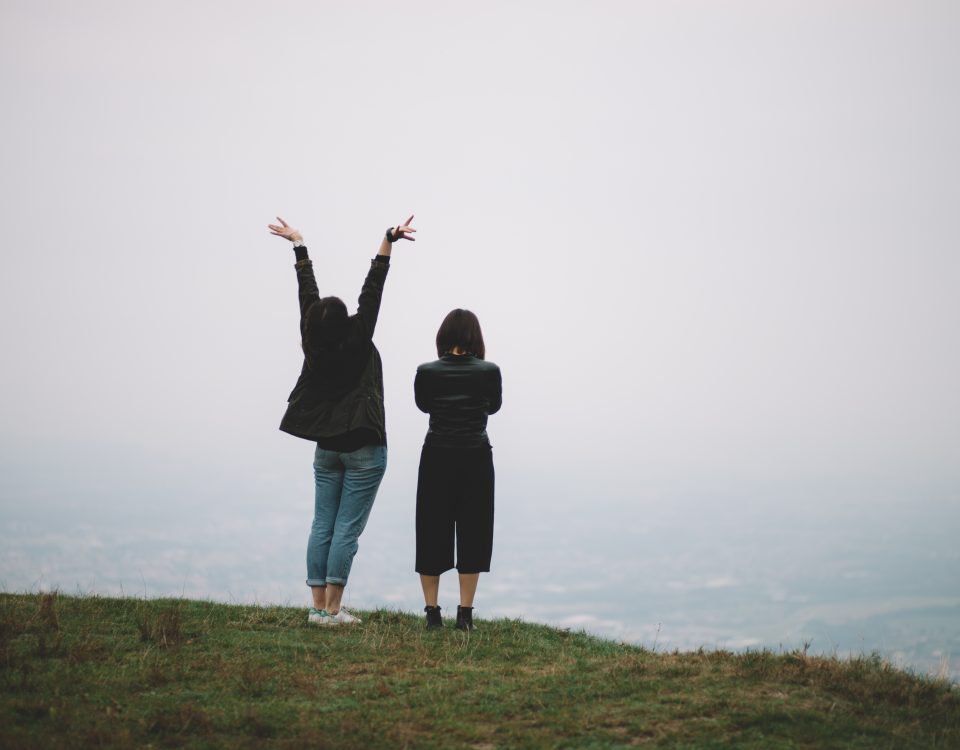 hiking vacation two women on a cliff edge looking out to see. one with folded arms and the other with arms in the air. dressed warmly in jeans and thick cardigans. grey sky, green grass foggy pale blue sea