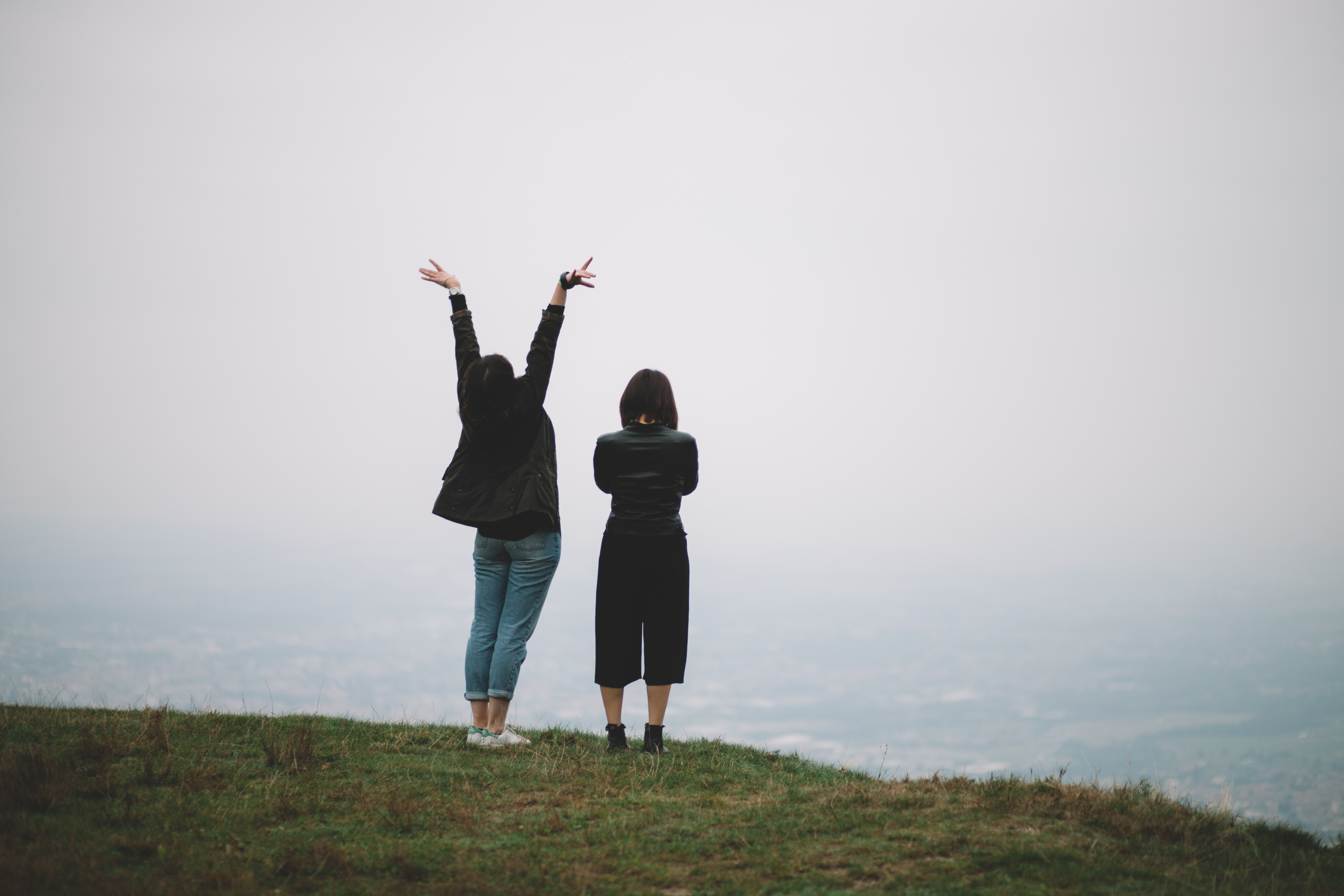 hiking vacation two women on a cliff edge looking out to see. one with folded arms and the other with arms in the air. dressed warmly in jeans and thick cardigans. grey sky, green grass foggy pale blue sea