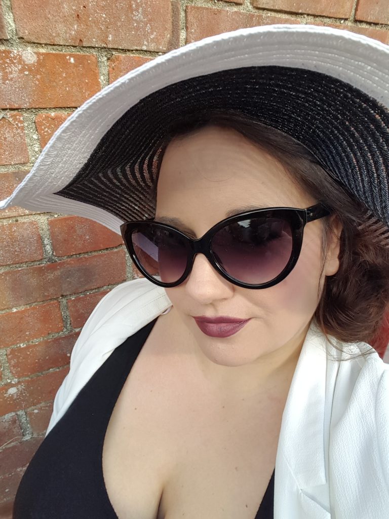 me wearing glasses a big hat and dark lipstick. Red wall in the background
