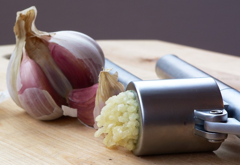 A bulb of garlic with cloves missing and crushed garlic coming out of a press all on a chopping board