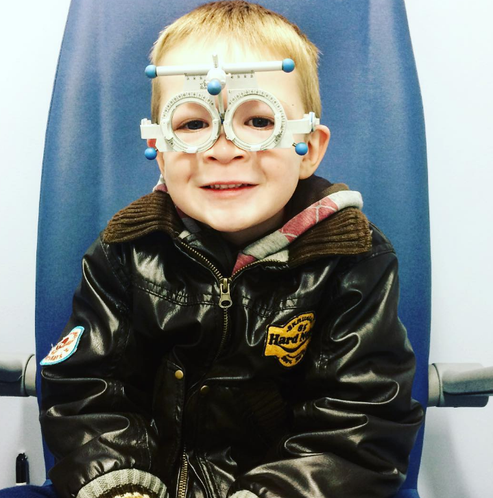 little boy smiling at the camera wearing a leath jacket with patches on and the tester glasses from an opticians sat in a blue chair