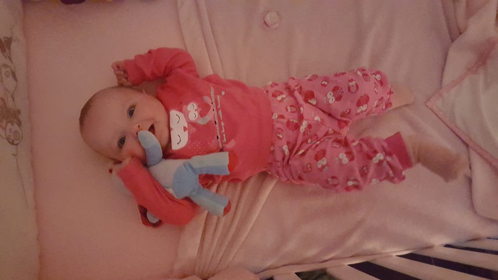 Alyssa at six months old lying in her cot in pyjamas cuddling iggle piggle smiling at the camera