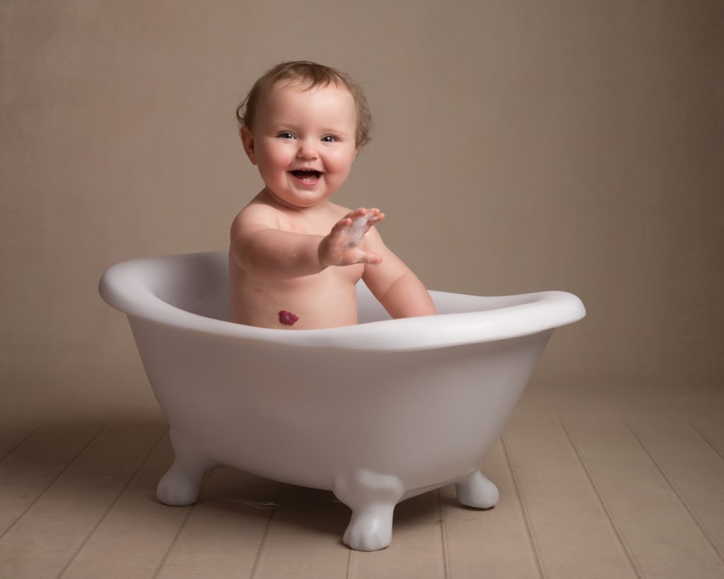 Alyssa sat in a mini free standing bath smiling at the camera holding out her hand with bubbles on the end