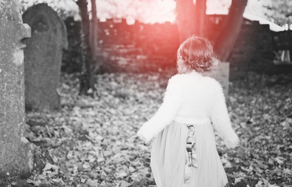 little girl dressed up in a dress and white jacket/cardigan walking away from the camera through the woods into pink light