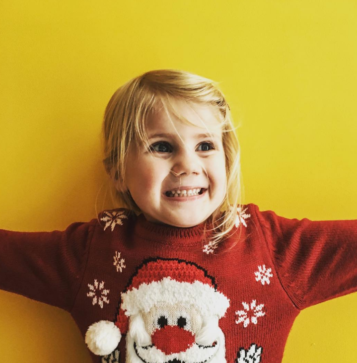 Blonde little girl smiling at something off camera wearing a red santa jumper in front of a yellow wall