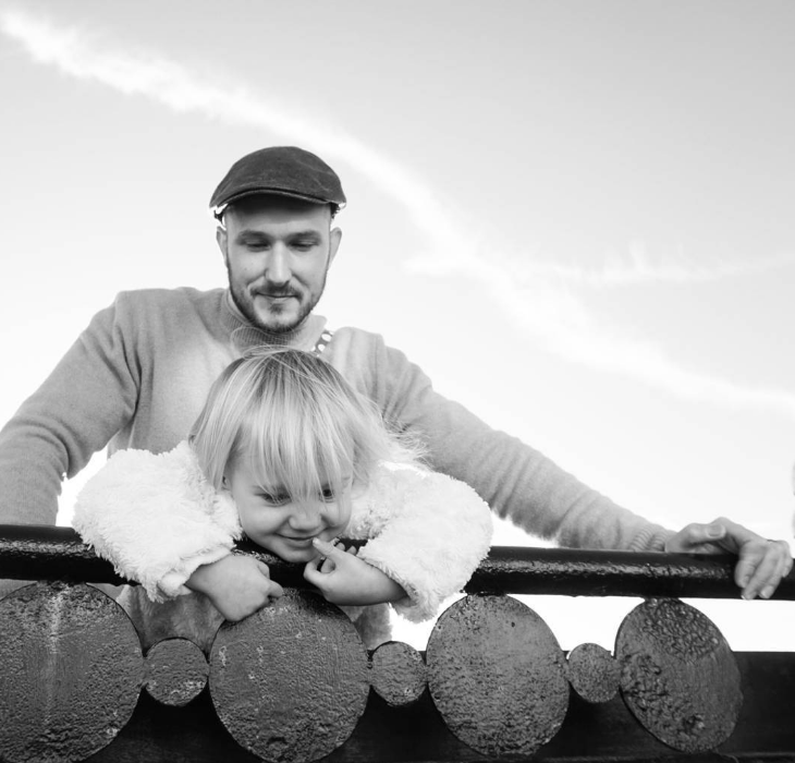 little blonde girl leaning over a wooden fence with her daddy above wearing a flat cap watching. picture in black and white