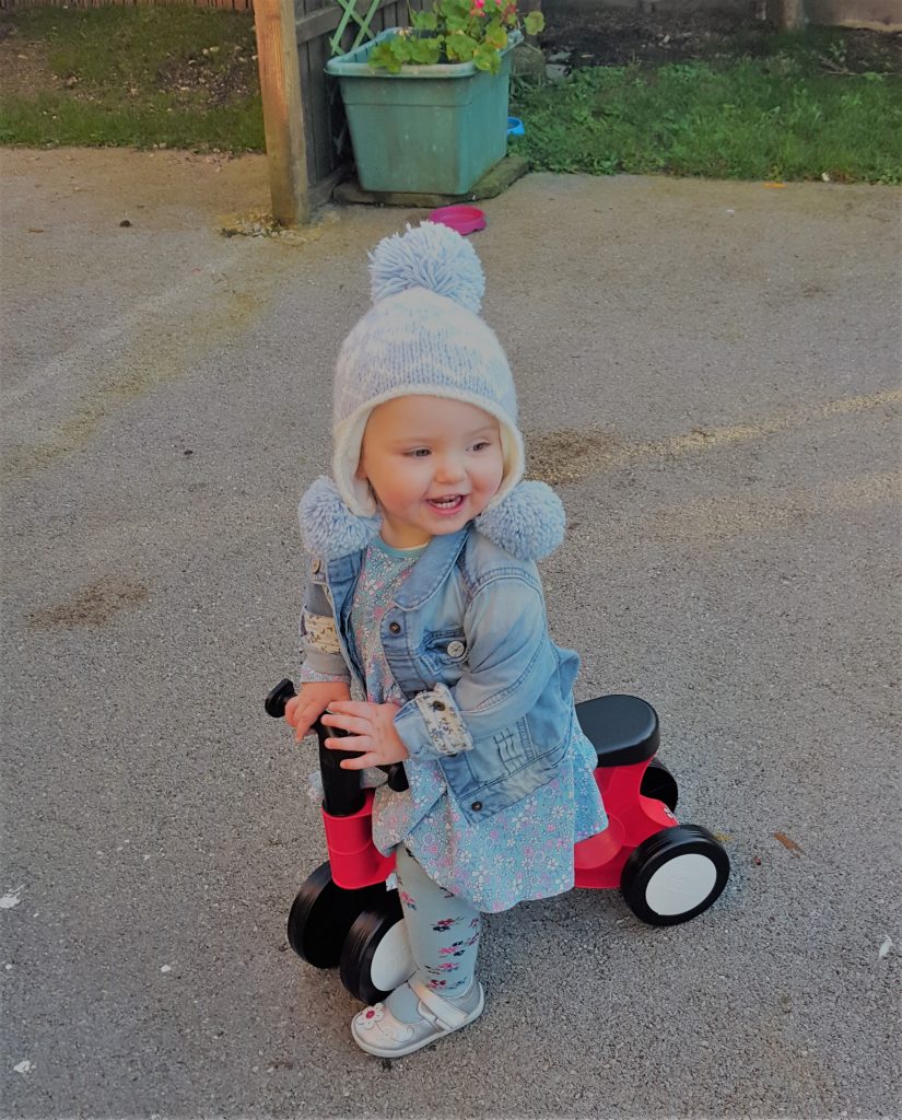 Alyssa in a wooly blue hat and denim jacket on a red toddler balance bike