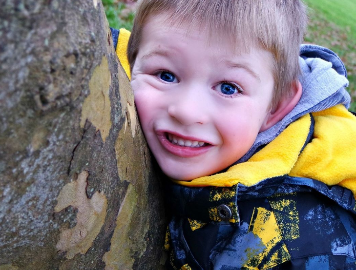 blonde little boy hugging a tree and smiling at the camera