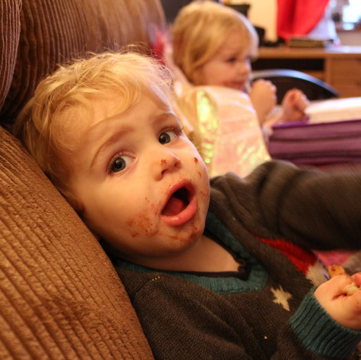 blonde haired boy looking at the camera with his mouth open and face covered in chocolate sat on brown sofa with blonde sibling in the background