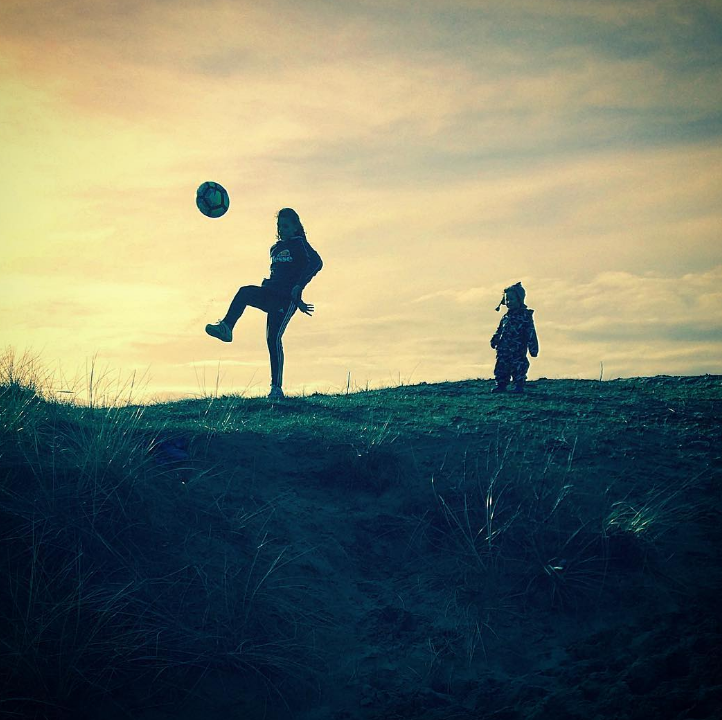 shadows of a girl kicking a ball in the air and a little toddler boy watching all on a grassy hill