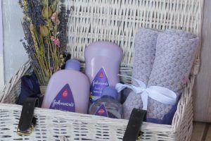 a white wicker hamper open with some dried flowers in a purple blacket tied with a white bow and 3 purple bottles of baby bath products - bubbles, wash and massage oil