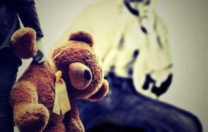a brown teddy hanging from a childs hand with a yellow ribbon round his neck