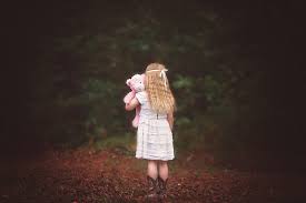 little blonde child facing away from camera standing in the autumn woods holding a pink teddy