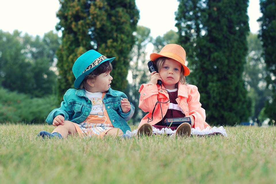 1 boy and 1 girl dress up in neone coloured suits over their clothes little girl looking at the camera on a play phone and little boy looking at her. alldone outside on grass with trees behind