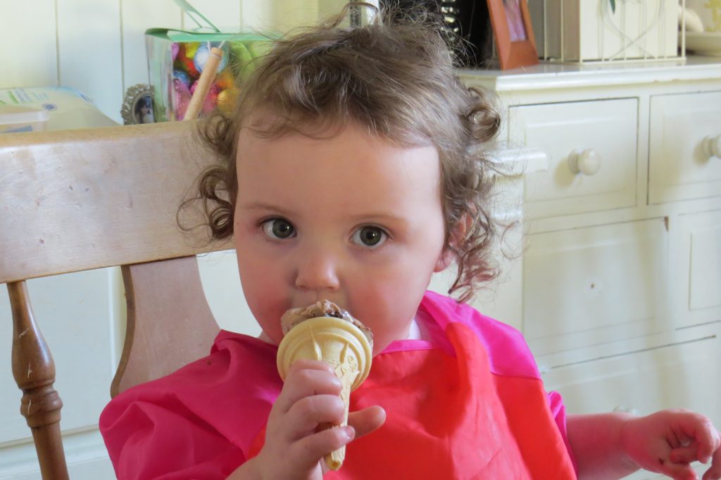 Alyssa in a red and pink apron eating a chocolate icecream cone with a beige sideboard behind her. she is looking at the camera