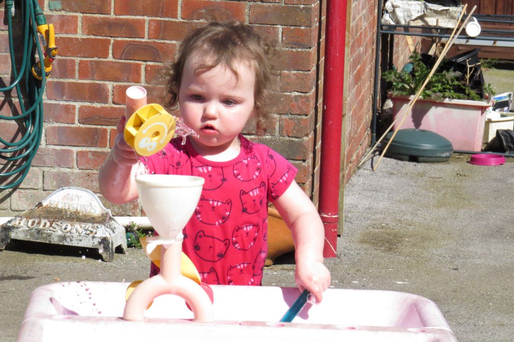 Alyssa in a pink t-shirt, in front of a brick wall playing at a water table with a funnel and a spoon