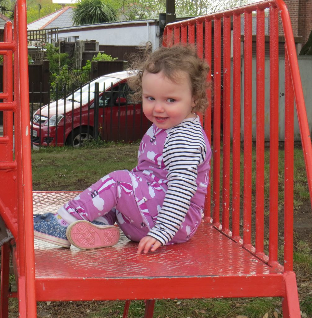 Alyssa sat on the red frame of a slide in a big black and white outfit with balloons on smiling at the camera with railings grass and a car in the background