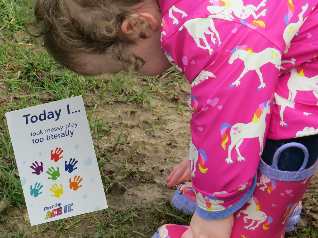 alyssa bending over to rub her hands in mud in her coat and wellies with a milestone card next to her saying that she has taken messy play to the next level