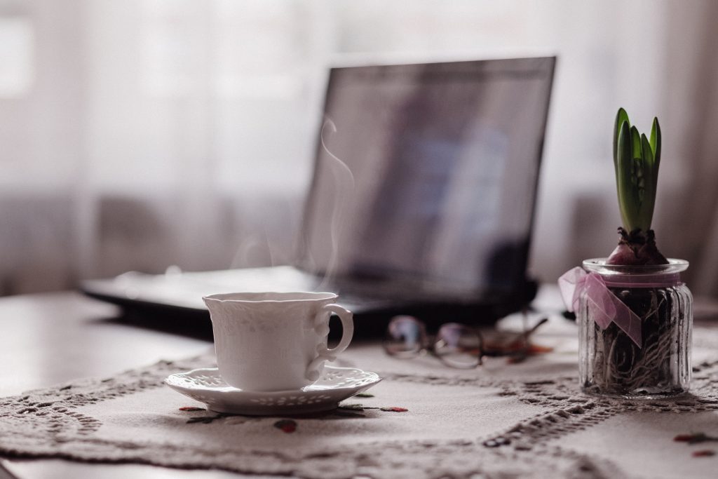 a laptop in the background open on a table. in front of it on a doily is a steaming cup of coffee in a white cup and saucer