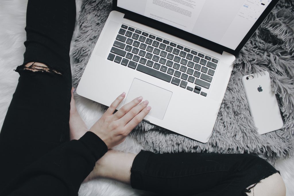 shot from above, a womans legs on a grey bed, wearing dark leggings, bare feet, with a silver laptop and one of her hands on the mouse pad