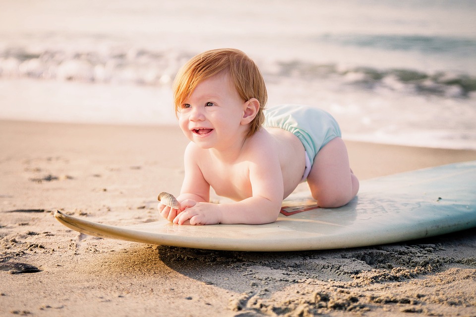 a red haired baby crawling ona white surfboard on the sand with a wave in the back ground and the sun to one side smiling at the camera