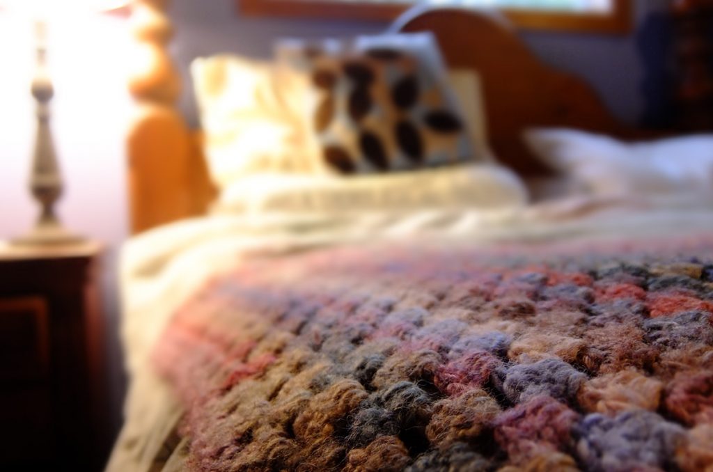 a crochet knit multi coloured blanket on a bed with a pillow and the bed head blurred in the background with a lamp to the left