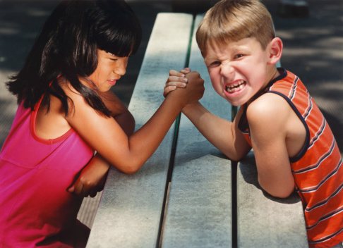 dark haired girl in pink top and fair haired boy in orange top gritting teeth and arm wrestling. boy looking at camera