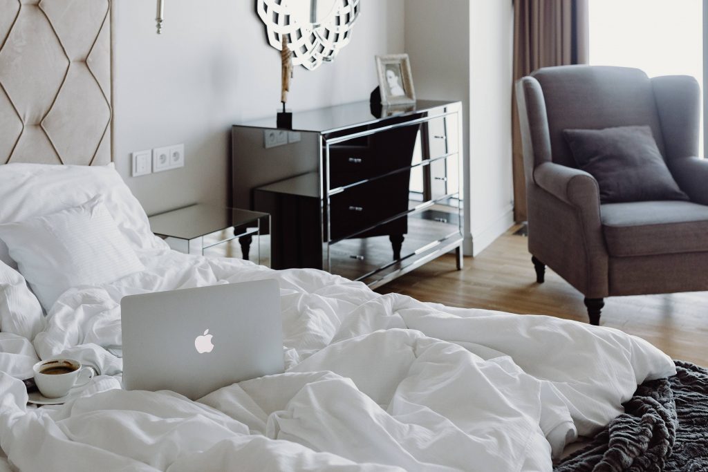 a white rumbled duvet on a bed with an apple mac silver laptop open a black reflective chest on the far wall with a couple of ornaments on top, wooden floor and a grey cuddle chair with a cushion in it