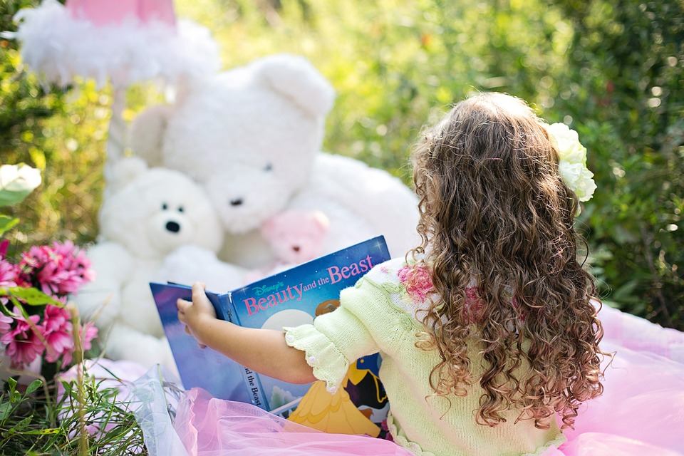 little girl back to camera with curly hair reading a book to teddy bears outside