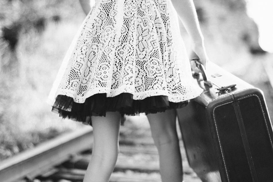 little girl in a lacy whitedress with underskirt can only be seen from waist down walking along a train track carrying a suit case. photo in black and white