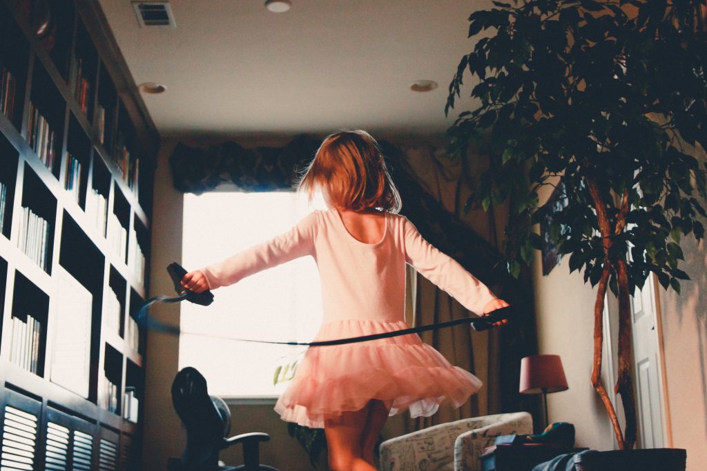young girl with her back to the camera in a hall way with bookshelves one side a window behind her and a plant on the right in a pink tutu and holding her arms out behind her with a skipping rope in them
