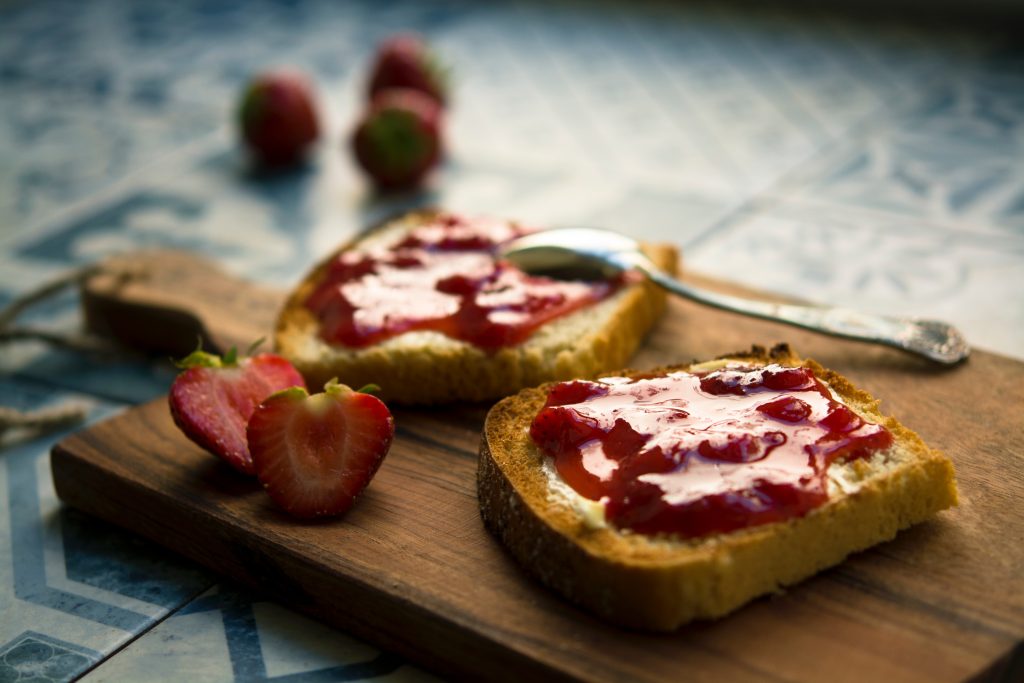 two slices of toast on a chopping board with a knife and strawberry jam on them and a strawberry cut in half on the board