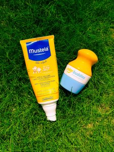a yellow and blue tube of Mustela suncream next to a yellow white and blue plastic solar buddies suncream applicator both on green grass
