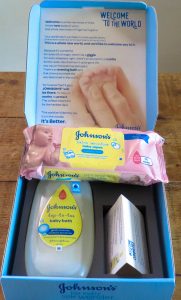 an open box on a wooden table with Johnsons wipes and body wash inside and an information leaflet. everything is pink blue and yellow