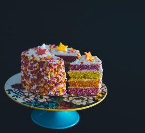 a 4 layered cake in pink yellow and orange seperated with white buttercream and covered on the outside in buttercream then multicoloured sprinkles and stars with a slice of the cake cut and pulled out slightly on a cake stand on a completely black background