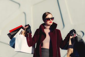 a woman in a red coat and sunglasses in front of a white building holding up lots of shopping bags in both hands