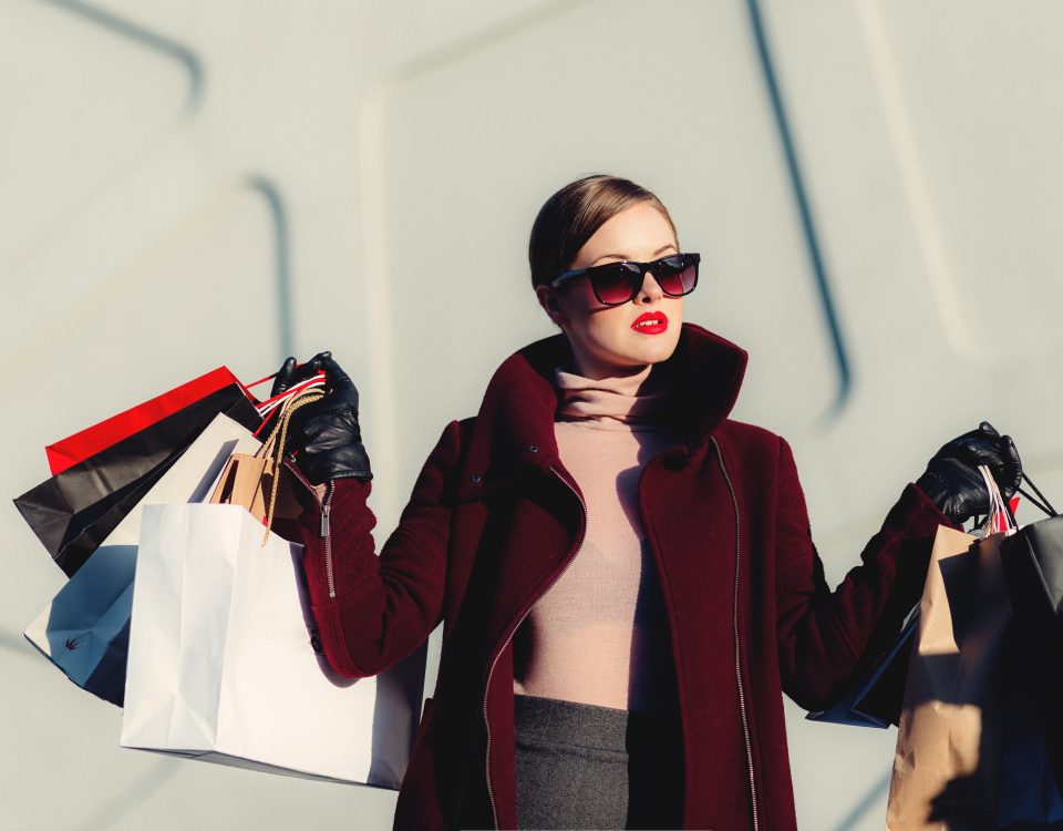 a woman in a red coat and sunglasses in front of a white building holding up lots of shopping bags in both hands