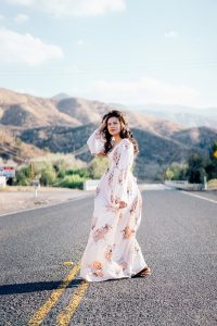 a woman wearing a white and floal maxi dress standing in the middle of a deserted mountain road pushing her brown hair out of her face to brighten your mood