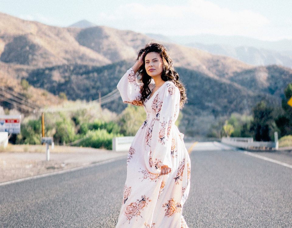 a woman wearing a white and floal maxi dress standing in the middle of a deserted mountain road pushing her brown hair out of her face