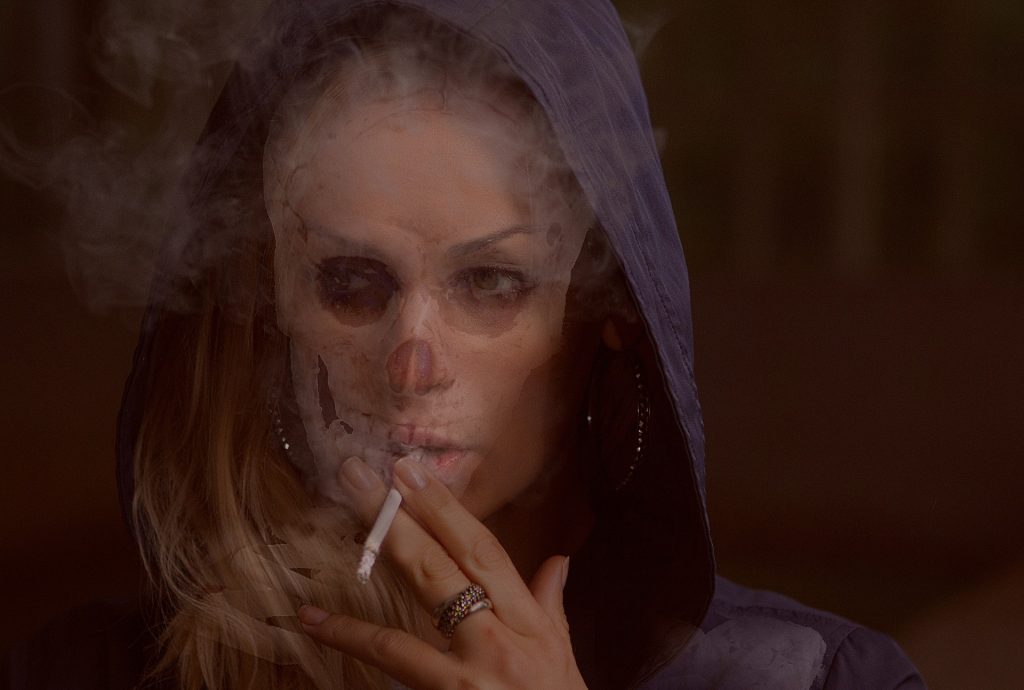 a woman in a blue hoodie with a cigarette to her mouth, smoke all around her and half her face appearing skull like