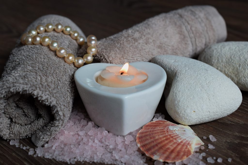 a purple towel with a heart shaped tea light candle lit, shells stones other rolled towels and pearls all around