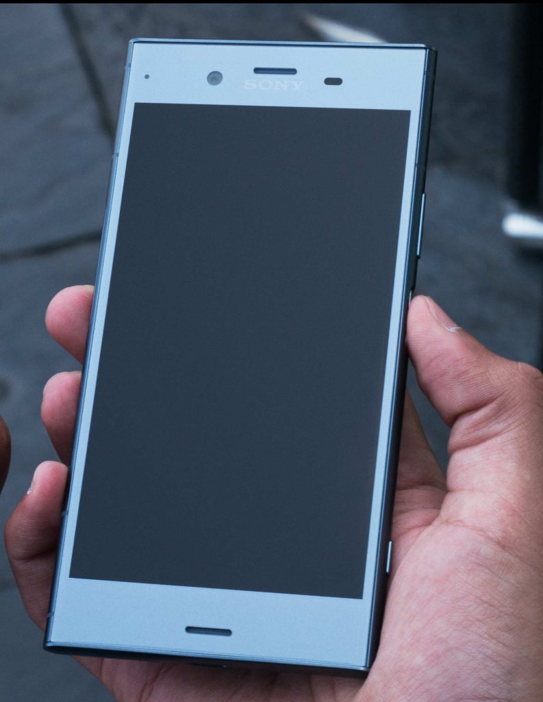 a hand holding a sony xperia xz1 compact in metallic blue screen up. the screen is black as the phone is off