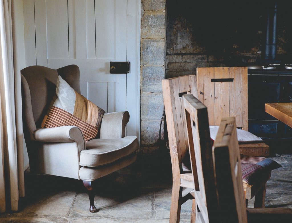 a wooden floor and bare brick walls a brown wing backed arm chair in the corner with a multi-tonal brown cushion on and to the right 4 wooden dining table chairs