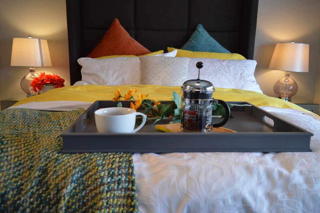 a photo from the bottom of a bed looking up. white bedlinen and orange and turquoise cushions with a black head board. yellow and green blanket. a breakfast tray with toast and coffee is on the end of the bed