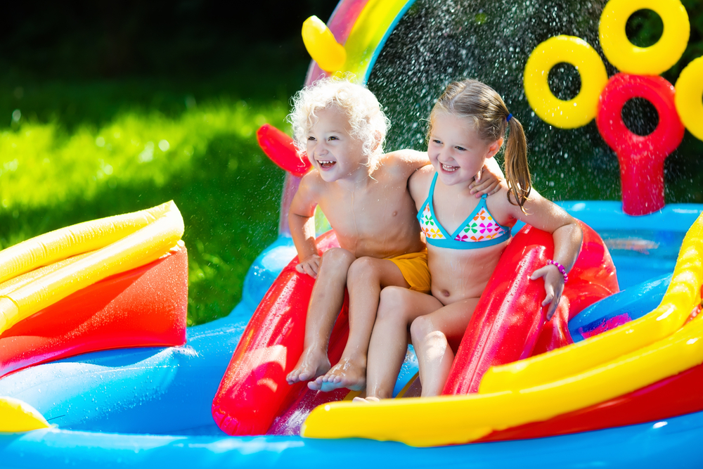 two children at the top of an inflatable slide in a paddling pool on a sunny day. costumes and pool red yellow and blue
