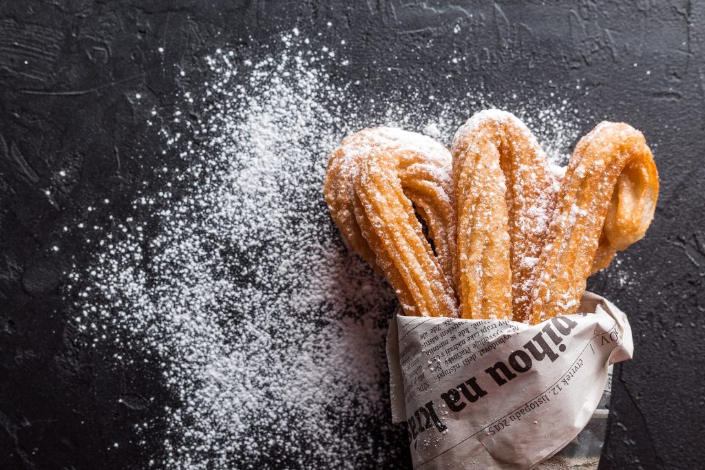 a black slate with icing sugar on it and a paper packet with 3 churros sticking out of it