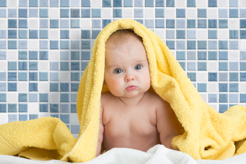 a baby sat on a white towel naked looking at the camera with a yellow towel over its head and blue tiles behind it
