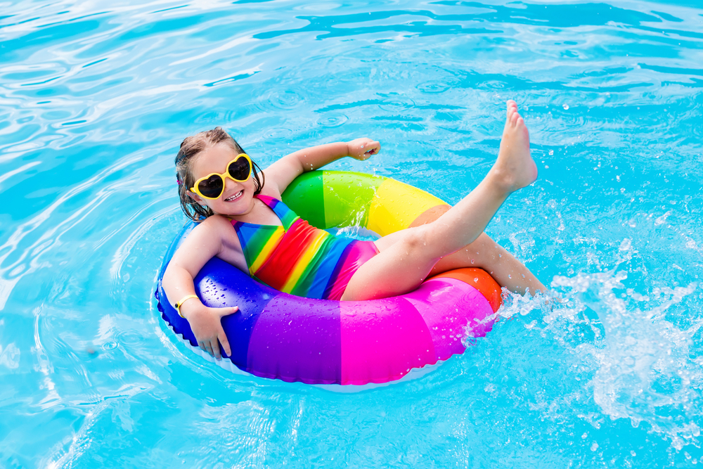 ayoung girl wearing sunglasses and a rainbow swimming costume floating in a rainbow rubber ring on a pool