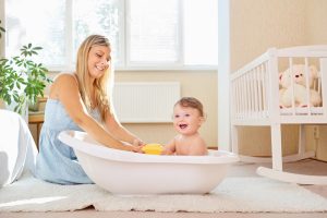 a mother bathing her baby in a yellow room in a white baby bath on the floor. both smiling at camera. room sunny